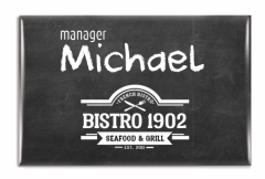 Rektangulär namnskylt med nål eller magnet. Till en bistro där bagrunden på namanbrickan ser ut som en dammig, svart tavla och texten gjorts så det liknar krita. Loggan i vitt nederst. Här har designen inspirerats av miljön där brickan ska användas.